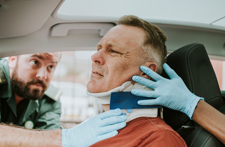A middle-aged man sits in a car wearing a neck brace, appearing to be in pain after a car accident. Two medical responders with blue gloves assist him—one adjusting the brace, the other providing support. Massaro Law Firm can help victims like him.