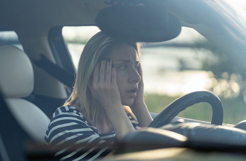 A woman wearing a striped shirt sits in a car, holding her head with both hands and looking distressed—perhaps after one of the DUI crashes that Massaro Law handles. Sunlight streams through the window, illuminating her worried face.