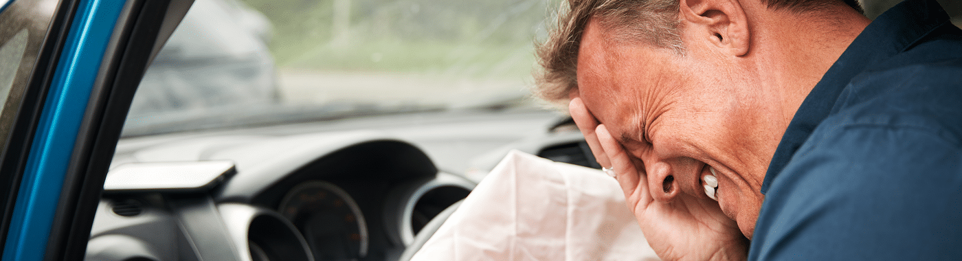 A man sits in a car, clutching his face in pain with a deployed airbag before him, suggesting he’s just been in a DUI crash—an incident Massaro Law can help you navigate.