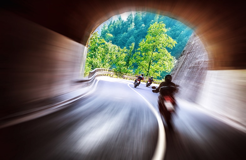 Three motorcyclists ride quickly out of a tunnel onto a winding forest road, motion blur suggesting speed and vibrant green trees in the sunlight outside—a scene where Motorcycle Accidents Massaro Law can help protect your rights if needed.