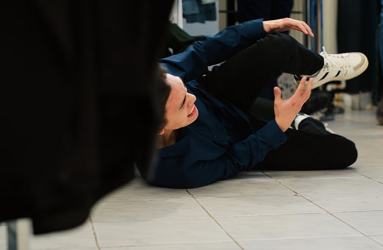 A person wearing a blue shirt, black pants, and white sneakers is lying on the floor indoors, appearing to be in pain and holding their leg after a slip and fall—an incident where Massaro Law can help.
