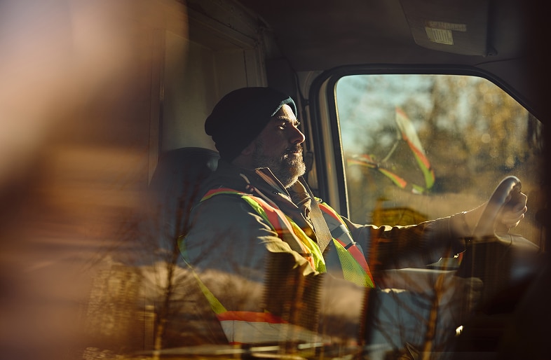 A man wearing a reflective safety vest and beanie drives a vehicle, with sunlight and trees reflected on the window, evoking the layered reality often seen in trucking and commercial vehicle accidents—areas handled by Massaro Law.