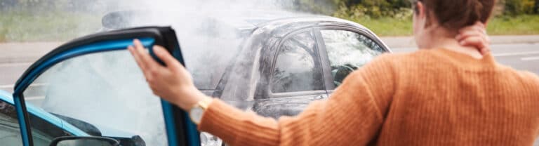 A person with a hand on their neck stands outside an open car door, looking at a black car with smoke coming from the hood after an apparent Uber or Lyft accident—perfect for a Delivery Crashes Featured Image.