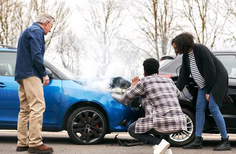 Three people inspect damage to two cars after a collision—one blue with smoke coming from its hood, the other black. The scene, outdoors on a cloudy day, highlights vehicle property damage and could involve diminished value claims. Massaro Law can help.