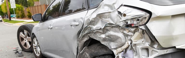 A silver car with severe rear-side damage and a detached tire lies on a suburban street. This vehicle property damage may qualify for diminished value—contact Massaro Law for guidance. Trees and houses form the background of the accident scene.