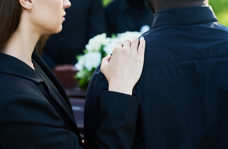 A woman in black gently rests her hand on a man's shoulder, offering comfort during a funeral; a casket and white flowers are visible in the background, reflecting the sorrow often seen in wrongful death cases at Massaro Law.