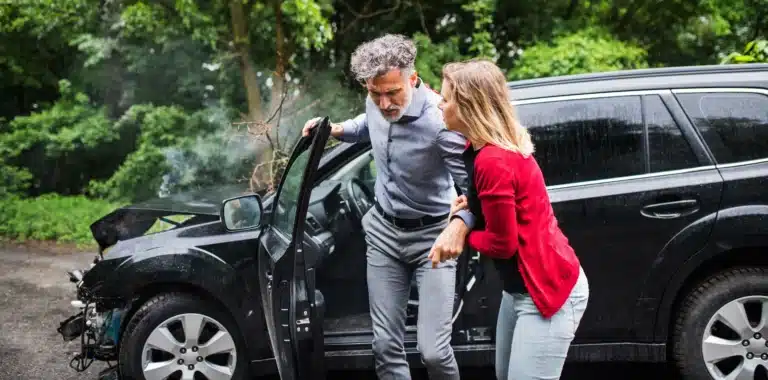 A woman helps an injured man out of a damaged black car after a car accident, with smoke rising from the front, in a wooded area—an incident where guidance from Massaro Law Firm could be crucial.