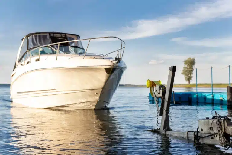 A white motorboat partially in the water near a dock, with a boat trailer and winch in the foreground. The sky is blue with scattered clouds, and there’s a floating platform and tree in the distance—a reminder to avoid boating accidents; Massaro Law can help.