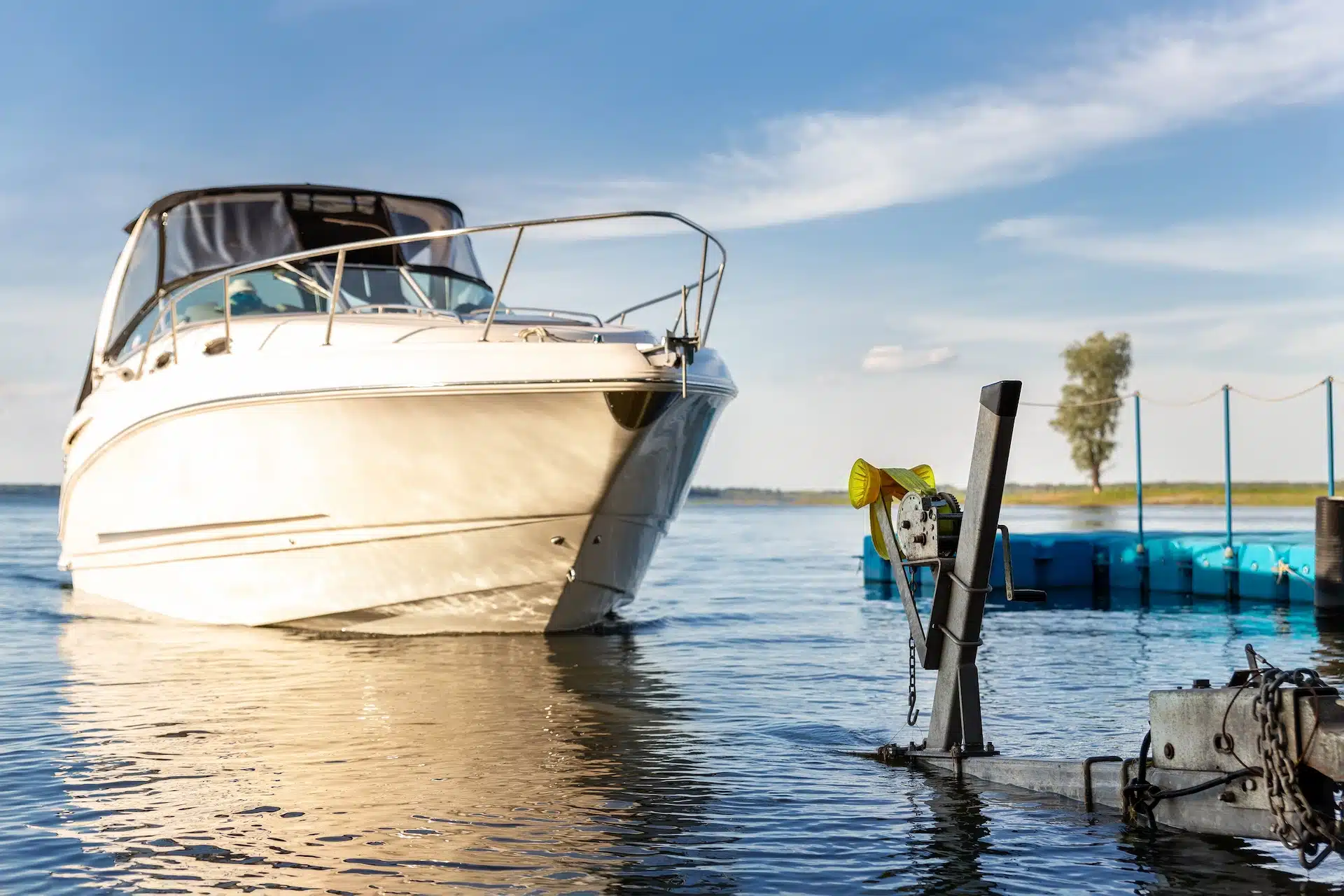A white motorboat partially in the water near a dock, with a boat trailer and winch in the foreground. The sky is blue with scattered clouds, and there’s a floating platform and tree in the distance—a reminder to avoid boating accidents; Massaro Law can help.