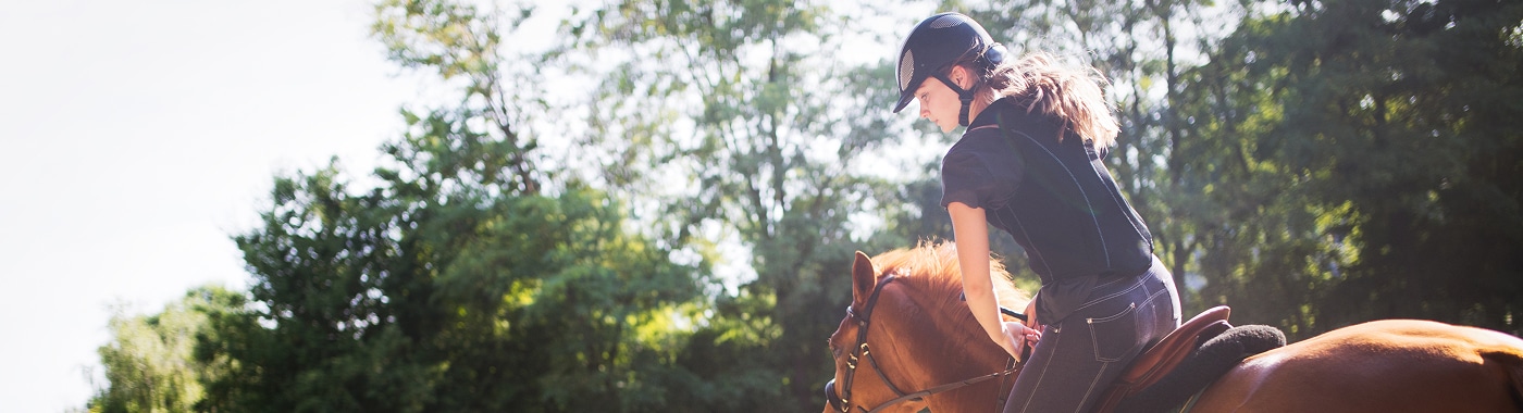 A person wearing a helmet and black shirt rides a brown horse outdoors on a sunny day, with green trees and bright sunlight in the background—a reminder to stay safe and informed about equine accidents with Massaro Law.
