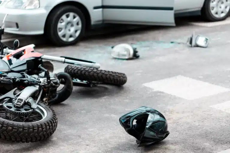 A motorcycle and helmet lie on the road near broken glass and car parts, next to a silver car with an open door—an all-too-common scene in Motorcycle Accidents Massaro Law handles at crosswalks.
