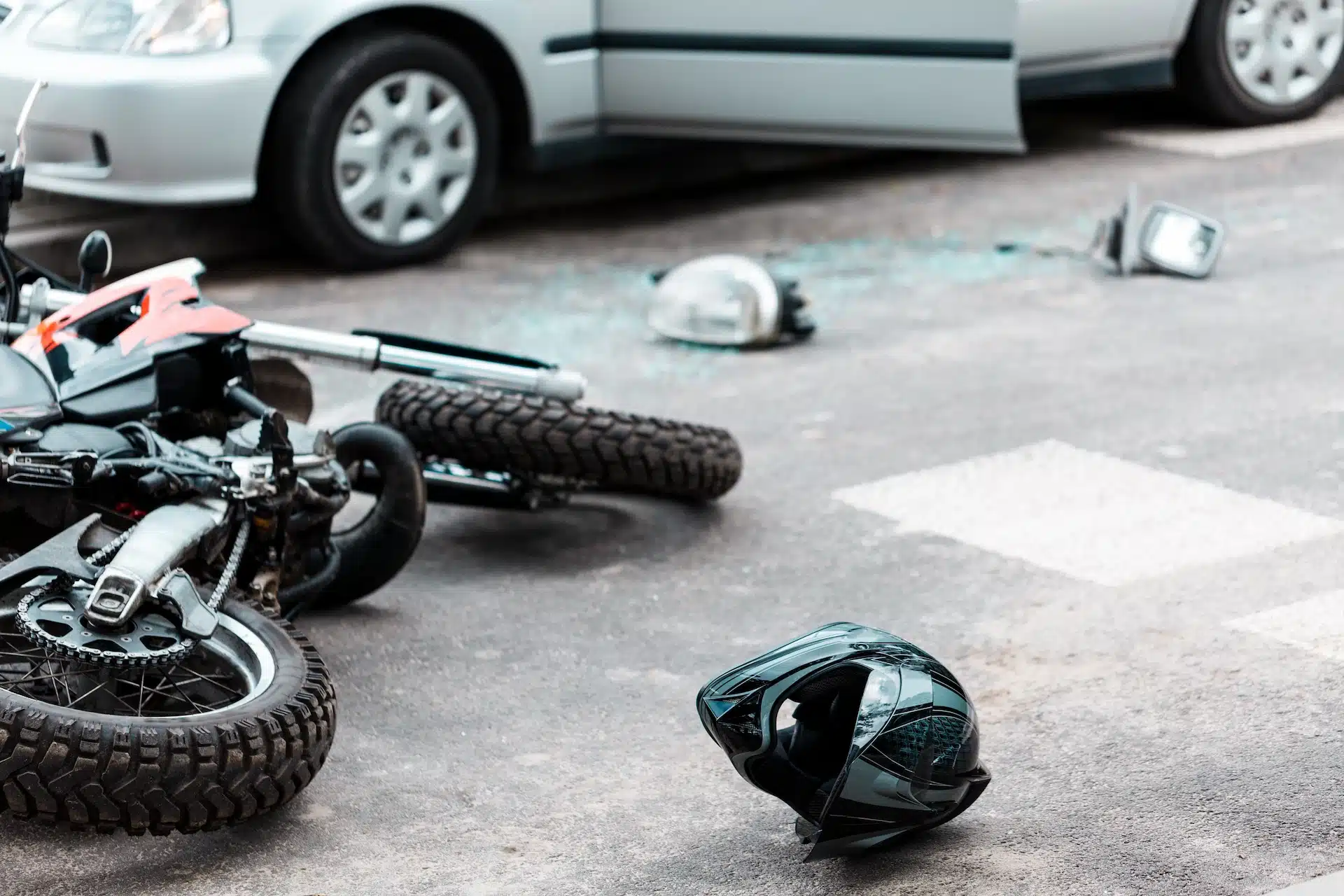 A motorcycle and helmet lie on the road near broken glass and car parts, next to a silver car with an open door—an all-too-common scene in Motorcycle Accidents Massaro Law handles at crosswalks.