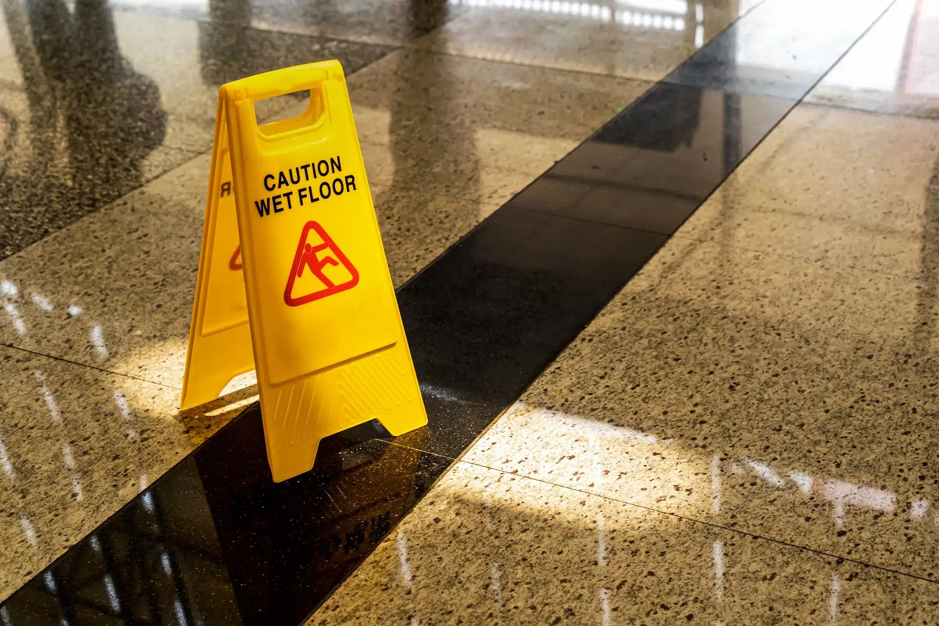 A yellow Caution Wet Floor sign stands on a shiny, reflective marble floor with sunlight streaming in and creating shadows—a reminder to prevent slip and fall accidents, as highlighted by Slip and Fall Massaro Law.