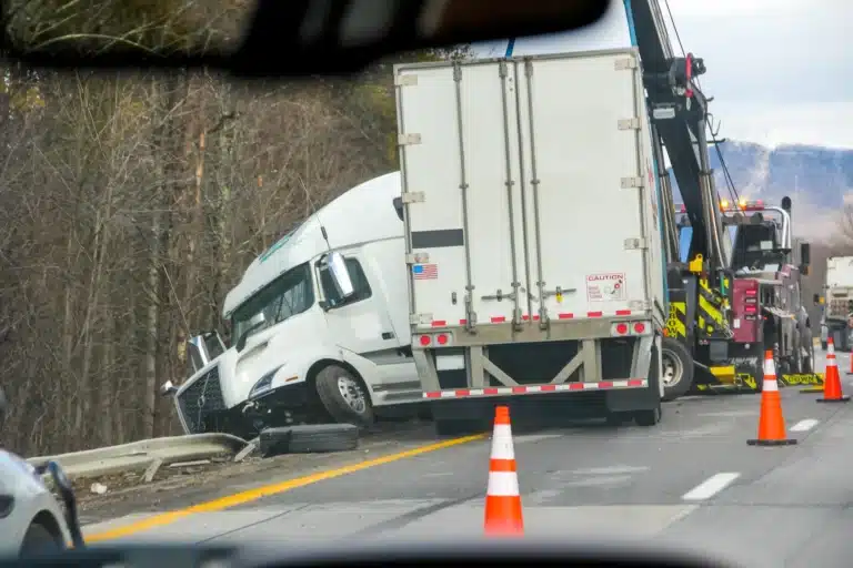 A white semi-truck crashed into a guardrail on the highway, with a tow truck and orange traffic cones nearby. Trees line the road as emergency response addresses the scene—Trucking and Commercial Vehicle Accidents Massaro Law Firm can help.