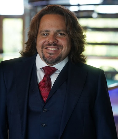 Vincent Massaro of Massaro Law Firm, with shoulder-length brown hair and a goatee, smiles while wearing a dark suit, white shirt, and red tie, standing indoors with soft, natural light in the background.