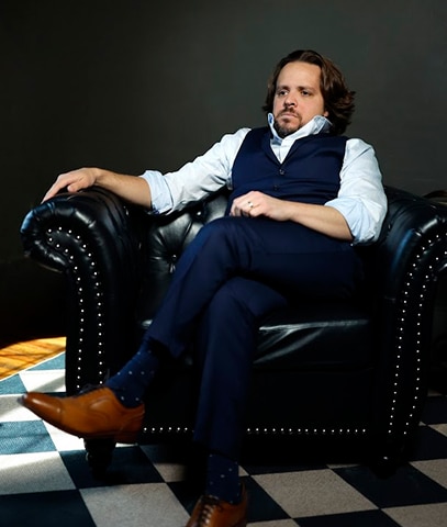 Vincent Massaro, a seasoned trial lawyer and founder of Massaro Law, sits relaxed on a black leather armchair with silver studs, dressed in a navy blue suit and brown shoes, on a black-and-white checkered floor in a dimly lit room.