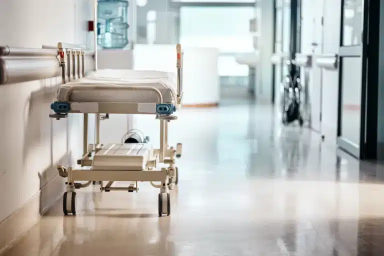 An empty hospital gurney is parked in a clean, bright hallway with a water cooler and wheelchair visible in the background, reflecting the calm environment where Wrongful Death Massaro Law Firm may review critical cases.